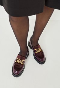 Loaferi din piele lăcuită burgundy, cu accente texturate, detalii din metal auriu și talpă groasă neagră, asortați cu ciorapi negri transparenți.