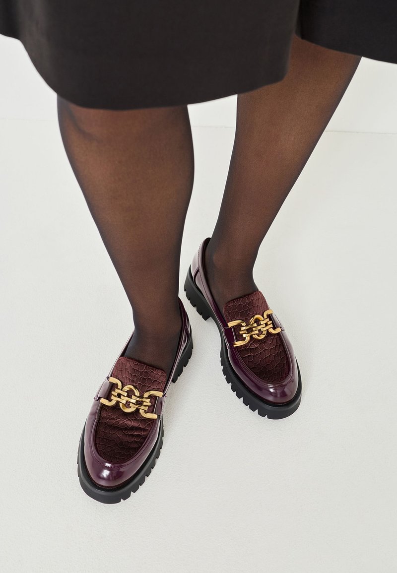 Loaferi din piele lăcuită burgundy, cu accente texturate, detalii din metal auriu și talpă groasă neagră, asortați cu ciorapi negri transparenți.