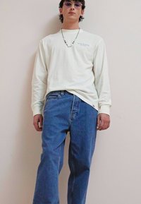Long-sleeve ivory shirt with blue logo, paired with loose blue jeans. Accessories include purple sunglasses and a beaded necklace.