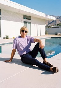 Jeune homme portant des lunettes de soleil, un t-shirt violet clair et un pantalon foncé, assis près d'une piscine moderne à côté d'un bâtiment blanc par une journée ensoleillée.
