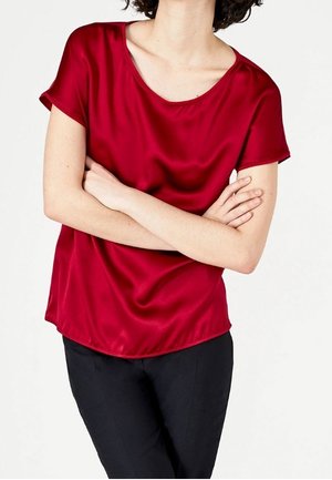Personne portant un chemisier en satin rouge à manches courtes et un pantalon noir, les bras croisés, debout devant un fond blanc uni.
