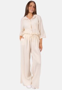 Zweiteiliges, hellbeiges Outfit aus strukturiertem Stoff. Das Oberteil hat einen Knopfverschluss mit einer Brusttasche; die Hose hat einen elastischen Bund.