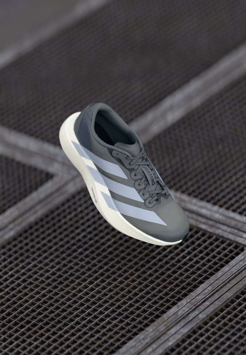 Zapatilla deportiva en gris y blanco, hecha de material sintético, con un diseño aerodinámico, acabado texturizado y un sistema de cordones prominente.