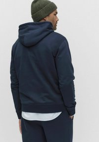 Mężczyzna w ciemnoniebieskiej bluzie z kapturem, granatowych spodniach i oliwkowej wełnianej czapce, stojący bokiem z rękami w kieszeniach na jednolitym tle.