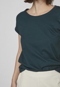 Dark teal short-sleeve t-shirt made of soft cotton, featuring a round neckline and relaxed fit, paired with light cream trousers.