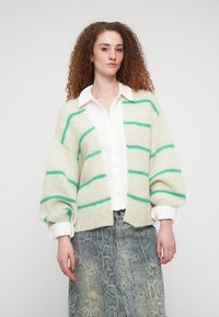 Πλεκτή ζακέτα σε ανοιχτό πράσινο με ρίγες σε τερρακότα, με υπερμεγέθη μανίκια και κοντή εφαρμογή, φορεμένη πάνω από ένα λευκό πουκάμισο με κουμπιά και μανίκια γυρισμένα.