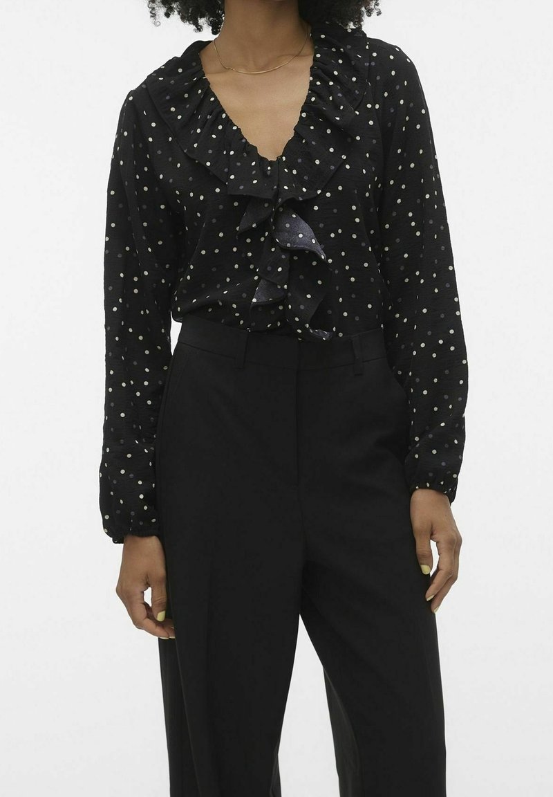 Schwarze Bluse mit weißen Punkten, rüschenbesetztem Ausschnitt und Vorderseiten-Details. Lange Ärmel mit elastischen Bündchen. Kombiniert mit schwarzen, taillierten Hosen.