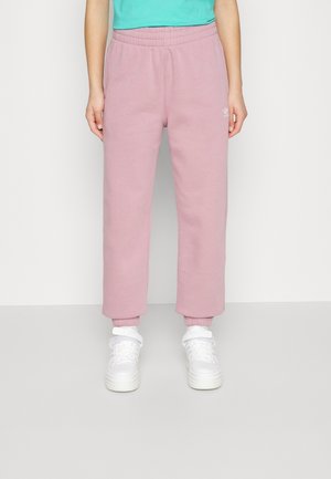 Person iført lyserøde Adidas joggingbukser og hvide sneakers står mod en ensfarvet lys baggrund, vist fra taljen til fødderne.