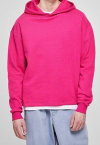 Felpa con cappuccio di un brillante colore rosa, dal taglio morbido, con polsini a costine e una texture liscia. Indossata sopra una maglietta bianca, presenta un design semplice.