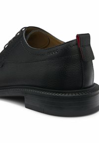 Schwarzer Textilleder-Herrenschuh mit Schnürung vorne, gestapelter Sohle und rotem Akzent am Fersenlaschenpuller, seitlich mit "Hugo" gebrannt.