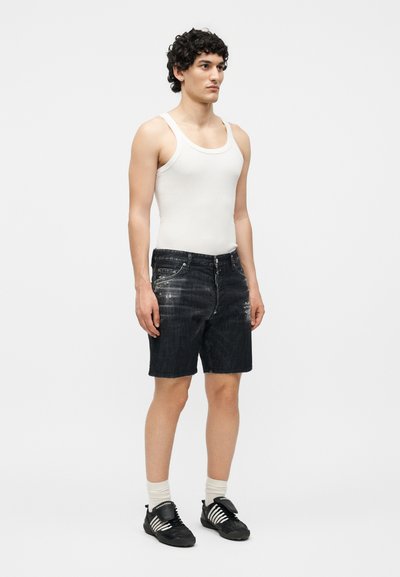 Άνδρας με σγουρά μαλλιά που φοράει λευκή φανέλα, μαύρα φθαρμένα τζιν σορτς, λευκές κάλτσες και μαύρα αθλητικά με άσπρες ρίγες, όρθιος.