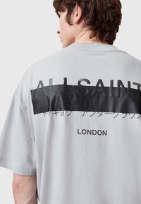 Camiseta gris de manga corta con un panel negro que presenta el texto en relieve "ALLSAINTS" y "LONDRES" debajo. Material de algodón suave, corte relajado.