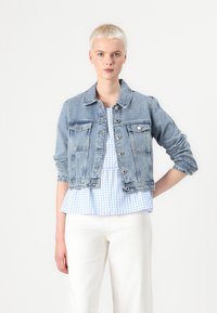 Chaqueta de denim azul claro con cierre de botones, dos bolsillos en el pecho y mangas enrolladas, superpuesta a una blusa peplum de cuadros vichy azul claro.