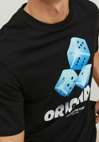 Svart bomull t-shirt med en grafik av tre blå tärningar med svarta prickar, och ordet "ORIGINAL" i fet vit text nedanför.