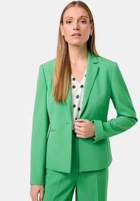 Grüner taillierter Blazer und passende Hose, aus glattem Stoff, mit Reverskragen und strukturierter Silhouette, kombiniert mit einer gemusterten weißen Bluse.