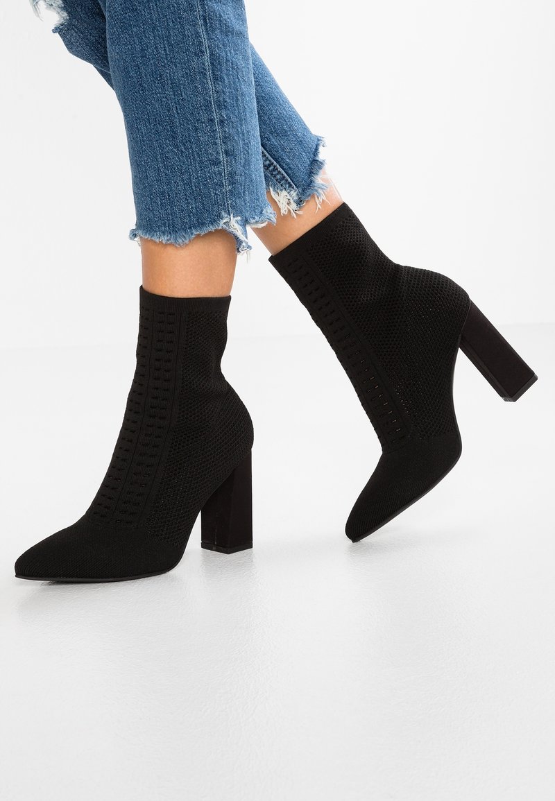 Jambes portant un jean bleu effiloché et des bottines en maille noire à talons carrés sur un sol blanc réfléchissant.