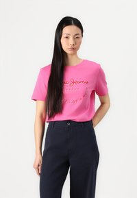 Camiseta de manga curta rosa-choque com texto bordado. Usada com calças azuis-marinho, a modelo está em pé contra um fundo branco liso.