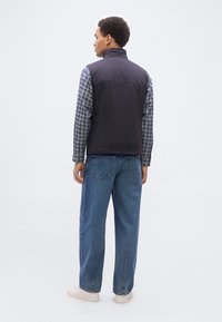 Άνδρας με μαύρο αμάνικο γιλέκο πάνω από μπλε καρό πουκάμισο, μπλε τζιν και λευκά αθλητικά παπούτσια, στέκεται γυρισμένος με την πλάτη σε ένα απλό φόντο.