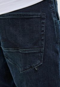 Jeans din denim închis au un buzunar posterior cu o cusătură curbată și o mică buclă; fabrică are o textură cu un ușor luciu și o culoare bogată.