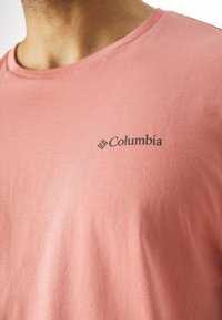 Kortärmad T-shirt med rund hals i ljusrosa bomull med en mjuk textur och en liten svart Columbia-logotyp på vänster bröst.