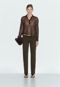 Chemise longue marron transparente à manches longues avec boutons et col, associée à un pantalon vert olive, rehaussée d'une ceinture cloutée et d'une pochette noire.