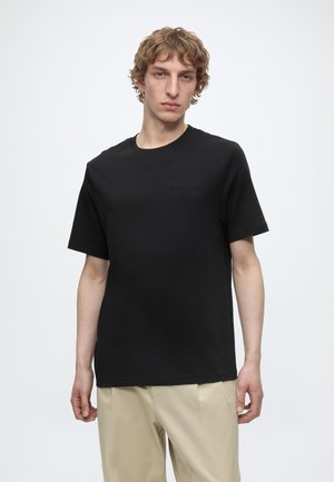 Jeune homme aux cheveux bouclés portant un t-shirt noir uni à manches courtes et un pantalon beige, debout devant un fond blanc uni.