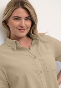 Chemise beige à boutons avec un col et une poche poitrine. Fabriquée en tissu texturé, à manches courtes, coupe décontractée.