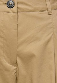 Gros plan sur la ceinture d'un pantalon beige avec un bouton noir à quatre trous et une passants de ceinture visible sur un tissu lisse.