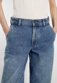 Das Modell trägt hochgeschnittene blaue Jeans mit einer entspannten Passform, die Hände in den Taschen, vor einem schlichten weißen Hintergrund.