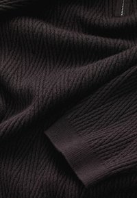 Donker textiel met een ribbelpatroon en een gladde manchette. De kleur is diepzwart, met subtiele variaties in de weving en glans.