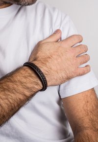 Brățară din piele împletită, de culoare maro închis, pe un încheietură, evidențiind o textură netedă și o formă circulară, pe un fundal de tricou alb.
