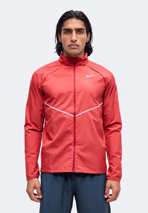 Mężczyzna z prostymi czarnymi włosami, ubrany w czerwoną kurtkę Nike na zamek z odblaskowymi akcentami i granatowe spodnie, stojący na tle jednolitego jasnego tła.