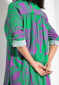 Faltiges Kleid mit grünem Grund und kräftigen lila Blumenmustern. Lange Ärmel mit aufgerollten Bündchen und einem lässigen Schnitt.