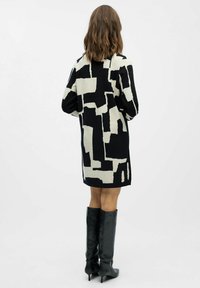 Strickkleid mit einem schwarz-creamfarbenen abstrakten Muster, lockerer Passform, langen Ärmeln und geradem Saum. Kombiniert mit kniehohen schwarzen Stiefeln.