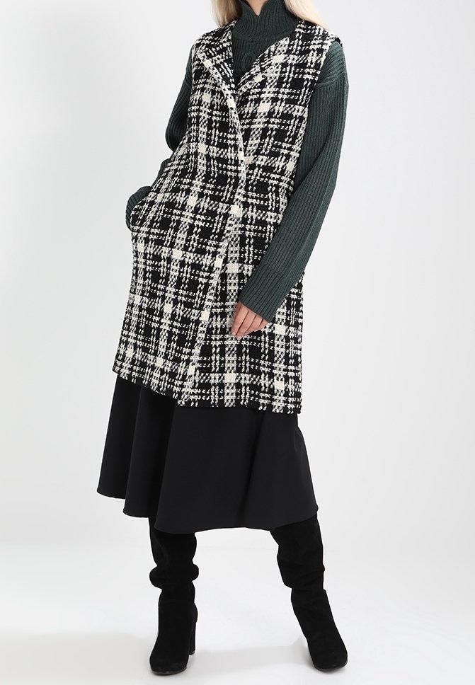 Kamizelka w kratę w czarno-białym kolorze z załamanymi klapami, teksturowanym materiałem, bocznymi kieszeniami, noszona na ciemnozielonym prążkowanym swetrze i czarnej spódnicy.