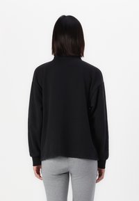 Svart sweatshirt med lange ermer, høy hals og avslappet passform. Myk tekstur, minimal sømdetalj ved hemmet. Utsikt fra baksiden.
