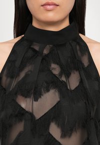Černý halter top z průhledného materiálu s vrstvenými třásněmi, strukturovanými vzory a hladkým, vysokým výstřihem.