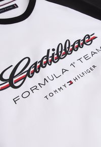 Bela majica s črnim ovratnikom z napisom "Cadillac Formula 1 Team" in logotipom Tommy Hilfiger s rdečimi, belimi in modrimi črtami.