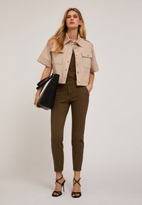 Chaqueta de color beige corta con mangas cortas y grandes bolsillos frontales, combinada con pantalones ajustados de color verde oliva y sandalias negras de tacón alto con tiras.