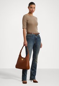 Bolsa de mão de couro castanho com forma curvada e detalhes em metal dourado, combinada com uma blusa justa bege e jeans flare azuis.