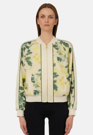 Donna che indossa una giacca bomber color crema con motivi floreali verdi e gialli, in piedi davanti a uno sfondo bianco semplice.