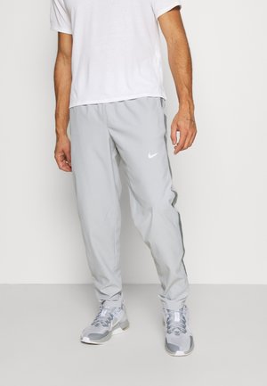 Mann trägt hellgraue Nike Trainingshose, weiße Nike und graue Sneakers, ein weißes T-Shirt und steht vor einem einfarbigen Hintergrund.