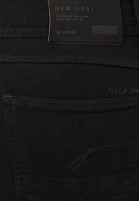 Vaqueros de mezclilla negra con una textura suave, que presentan un bolsillo trasero con costuras mínimas y una etiqueta de G-Star visible en la cinturilla.