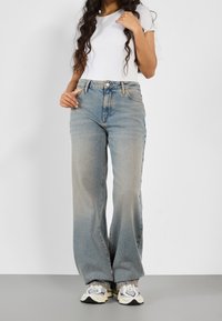 Jasnoniebieskie dżinsy typu wide-leg z przetartą fakturą, w połączeniu z dopasowanym białym t-shirtem i sneakersami z fioletowymi i granatowymi akcentami.