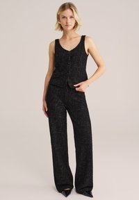 Črno tvid komplet, ki vključuje brezrokavni top z V-izrezom in gumbom, v kombinaciji s širokimi hlačami. Diskretni poudarki v večbarvni nitki.