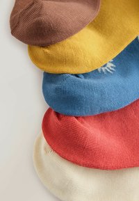 Pile de cinq bérets tricotés en marron, jaune moutarde, bleu avec motif blanc, rouge corail et crème sur une surface claire.