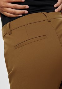Brown trousers featuring a flat back pocket, smooth texture, and tailored fit, with a visible waistband and side seam detail.