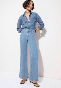 Denim-Outfit mit einem blauen Hemd mit Knöpfen, zwei Brusttaschen und hochtaillierten, hellblauen Schlagjeans mit seitlichen Nähten.