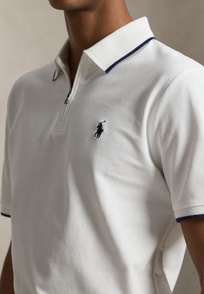 Homme portant un polo blanc avec un liseré bleu marine sur le col et les manches, et un logo brodé d’un joueur de polo bleu marine sur la poitrine.