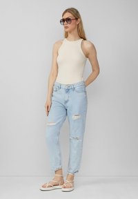 Crème ribbel tanktop, lichtblauwe high-waisted jeans met gescheurde knieën en witte sandaaltjes met bandjes. Model draagt oversized zonnebril.